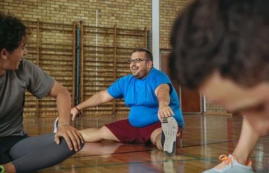 Ein Mann in blauem T-Shirt und roter Sporthose sitzt in einer Turnhalle beim Dehnen auf dem Boden, streckt ein Bein nach vorne und lächelt, während zwei weitere Personen unscharf im Vordergrund mittrainieren.
