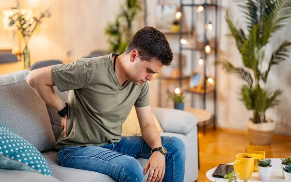 Ein junger Mann sitzt mit einer Hand an seinem schmerzenden Rücken auf dem Sofa, sein Blick geht nach unten.