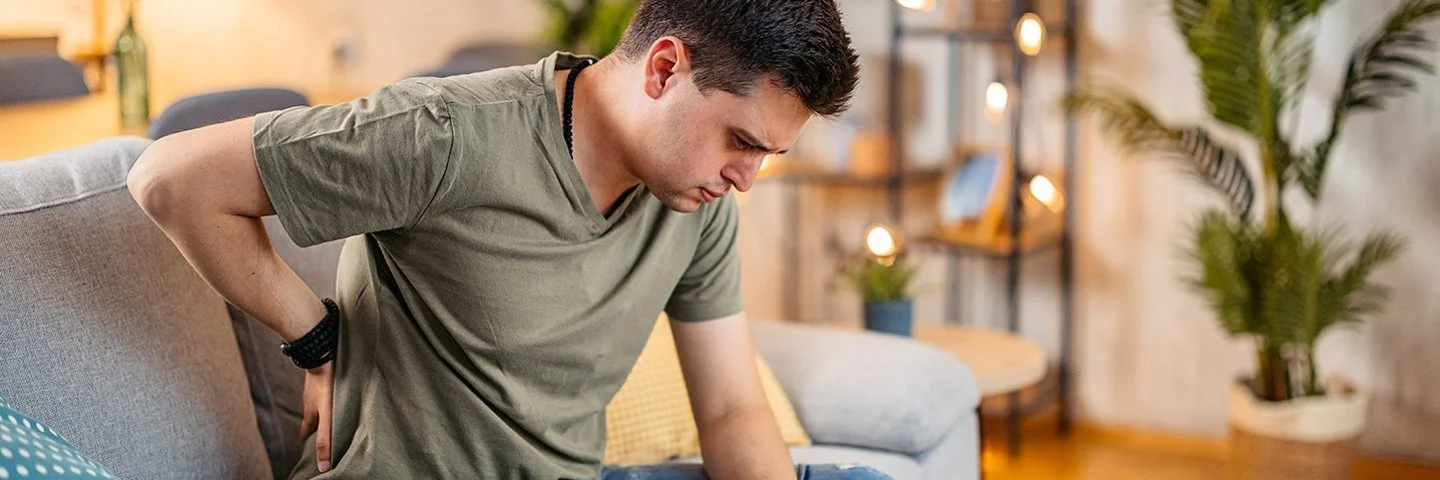 Ein junger Mann sitzt mit einer Hand an seinem schmerzenden Rücken auf dem Sofa, sein Blick geht nach unten.