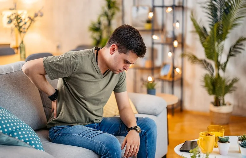 Ein junger Mann sitzt mit einer Hand an seinem schmerzenden Rücken auf dem Sofa, sein Blick geht nach unten.
