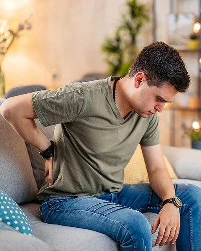 Ein junger Mann sitzt mit einer Hand an seinem schmerzenden Rücken auf dem Sofa, sein Blick geht nach unten.
