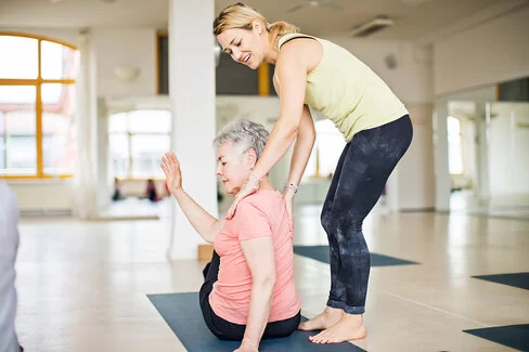 Eine Frau sitzt in einem Yogakurs auf ihrer Matte und führt eine Übung aus. Die Trainerin steht hinter ihr und leitet sie an.