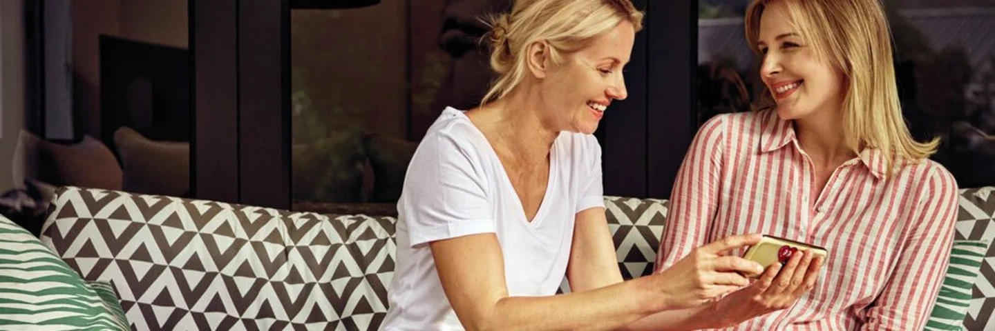 Zwei Frauen sitzen auf einer Couch mit gemusterten Kissen, eine zeigt der anderen etwas auf einem Smartphone.