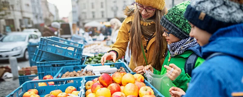 Drei Kinder mit Mützen kaufen regionale Äpfel auf einem Wochenmarkt.