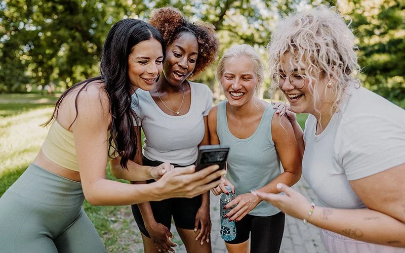 Vier Frauen in Sportkleidung stehen im Freien und schauen gemeinsam auf ein Smartphone.