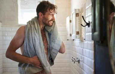 Ein unbekleideter Mann mit Handtuch steht lächelnd vor dem Spiegel im Badezimmer.