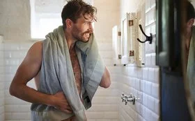 Ein unbekleideter Mann mit Handtuch steht lächelnd vor dem Spiegel im Badezimmer.