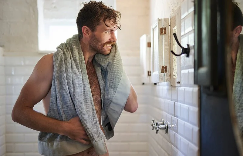 Ein unbekleideter Mann mit Handtuch steht lächelnd vor dem Spiegel im Badezimmer.