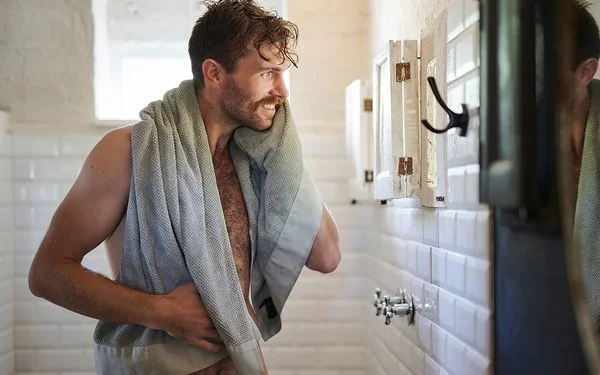 Ein unbekleideter Mann mit Handtuch steht lächelnd vor dem Spiegel im Badezimmer.