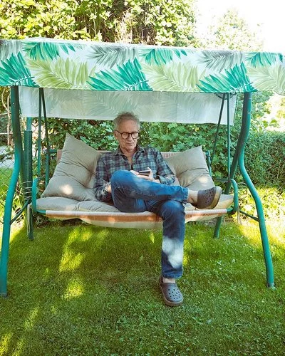 Ein älterer Mann mit Brille sitzt auf einer grünen Gartenschaukel mit grün-weiß gemustertem Sonnendach und schaut auf sein Handy.
