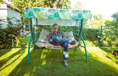 Ein älterer Mann mit Brille sitzt auf einer grünen Gartenschaukel mit grün-weiß gemustertem Sonnendach und schaut auf sein Handy.