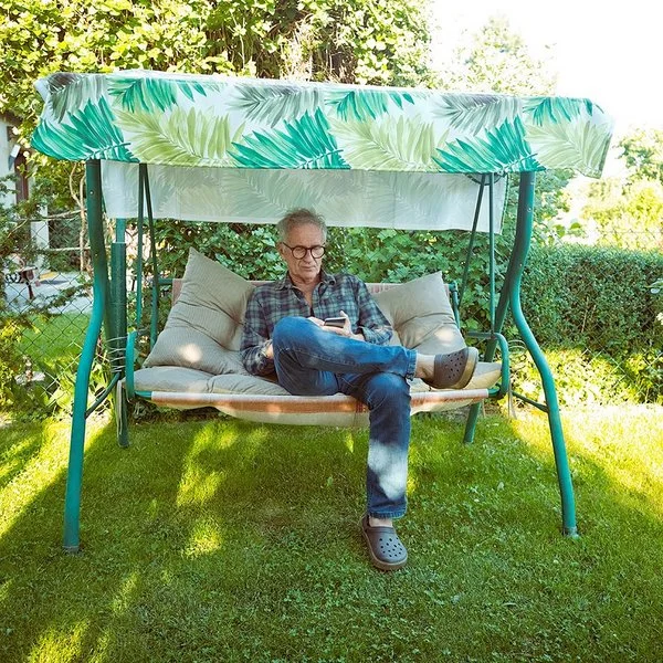 Ein älterer Mann mit Brille sitzt auf einer grünen Gartenschaukel mit grün-weiß gemustertem Sonnendach und schaut auf sein Handy.