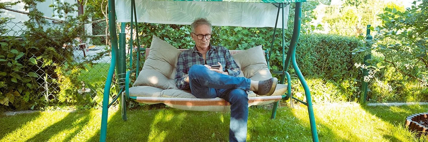 Ein älterer Mann mit Brille sitzt auf einer grünen Gartenschaukel mit grün-weiß gemustertem Sonnendach und schaut auf sein Handy.