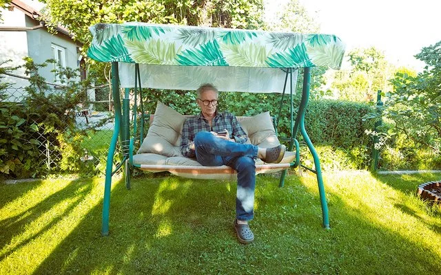 Ein älterer Mann mit Brille sitzt auf einer grünen Gartenschaukel mit grün-weiß gemustertem Sonnendach und schaut auf sein Handy.