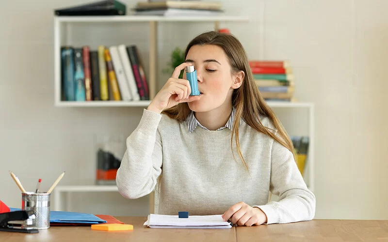 Eine junge Frau sitzt am Schreibtisch und benutzt einen Asthma-Inhalator.