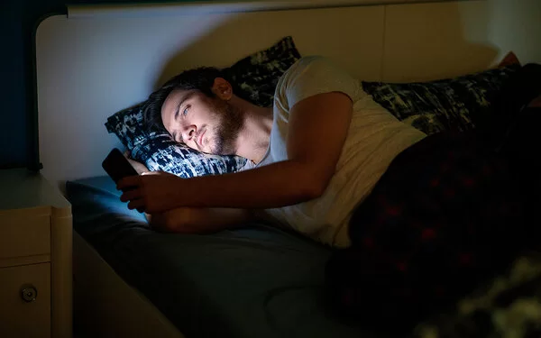Ein junger Mann liegt in seinem dunklen Schlafzimmer im Bett und schaut auf sein Smartphone.