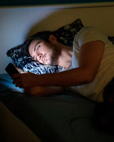 Ein junger Mann liegt in seinem dunklen Schlafzimmer im Bett und schaut auf sein Smartphone.