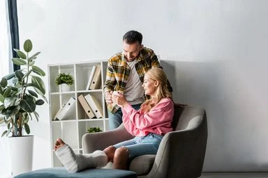 Eine Frau sitzt mit einem Gipsverband am Bein im Sessel, ein Mann reicht ihr ein Heißgetränk.