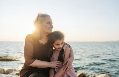 Eine etwa 60-jährige Frau hält ein etwa 9-jähriges Mädchen im Arm. Beide sitzen nebeneinander auf Steinen am Meer und schauen zur Seite.