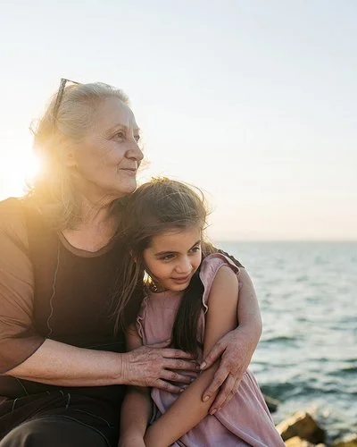 Eine etwa 60-jährige Frau hält ein etwa 9-jähriges Mädchen im Arm. Beide sitzen nebeneinander auf Steinen am Meer und schauen zur Seite.