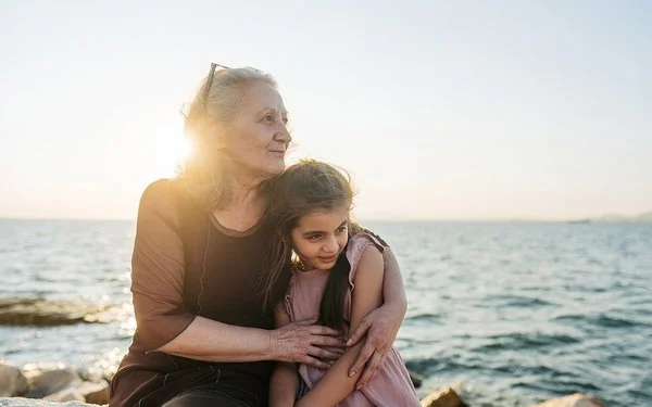 Eine etwa 60-jährige Frau hält ein etwa 9-jähriges Mädchen im Arm. Beide sitzen nebeneinander auf Steinen am Meer und schauen zur Seite.