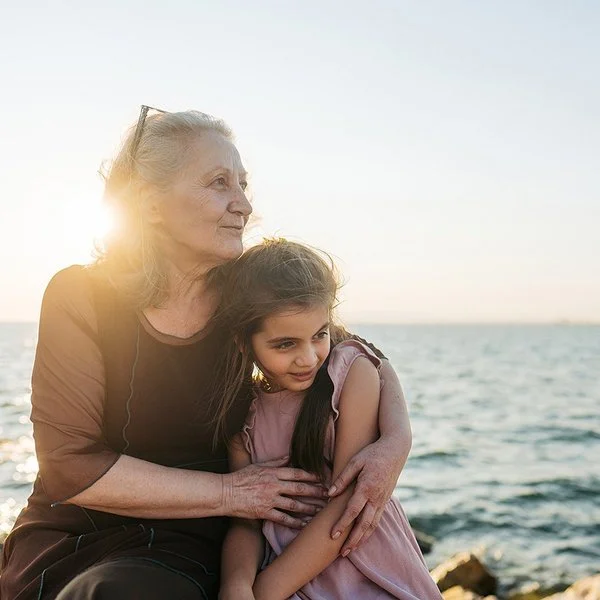 Eine etwa 60-jährige Frau hält ein etwa 9-jähriges Mädchen im Arm. Beide sitzen nebeneinander auf Steinen am Meer und schauen zur Seite.