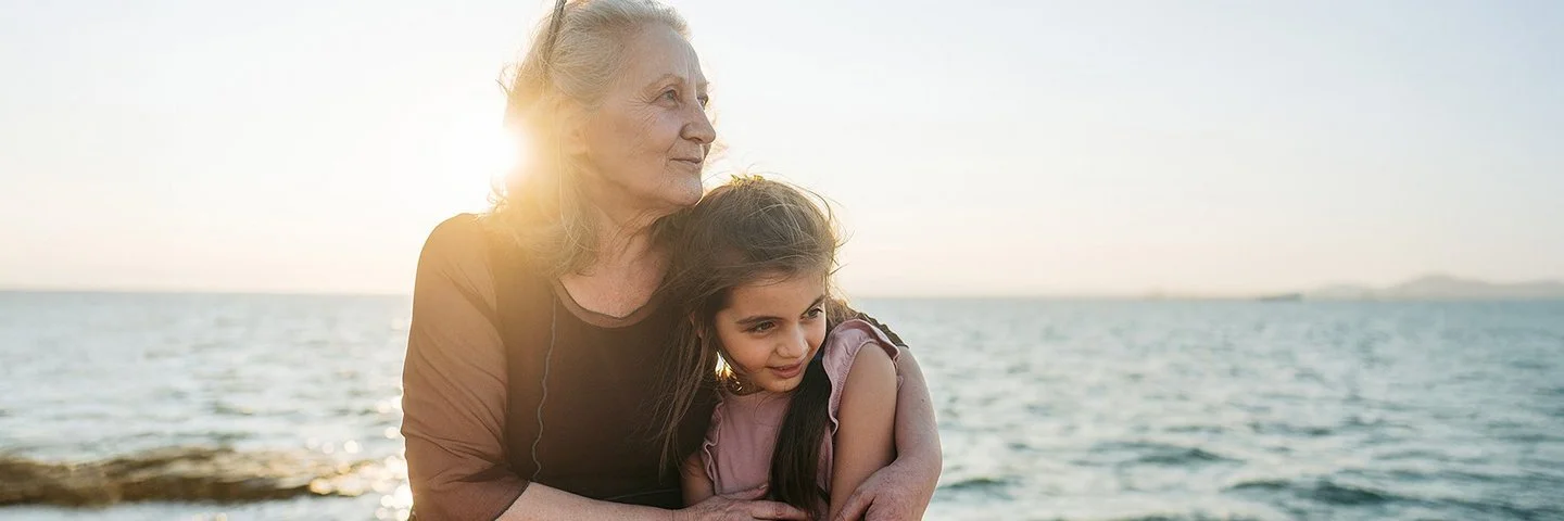 Eine etwa 60-jährige Frau hält ein etwa 9-jähriges Mädchen im Arm. Beide sitzen nebeneinander auf Steinen am Meer und schauen zur Seite.