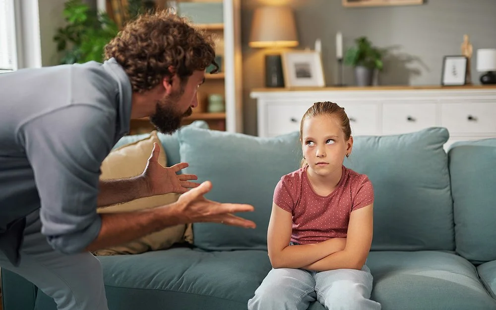 Ein Mann steht mit beiden Händen gestikulierend im Wohnzimmer vor einem auf dem Sofa sitzenden Mädchen. Das Mädchen hat die Arme vor dem Körper verschränkt und schaut ihm in die Augen.