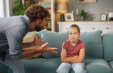 Ein Mann steht mit beiden Händen gestikulierend im Wohnzimmer vor einem auf dem Sofa sitzenden Mädchen. Das Mädchen hat die Arme vor dem Körper verschränkt und schaut ihm in die Augen.
