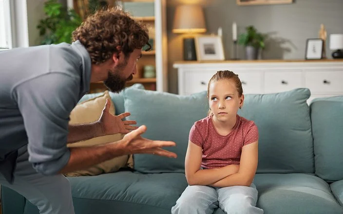 Ein Mann steht mit beiden Händen gestikulierend im Wohnzimmer vor einem auf dem Sofa sitzenden Mädchen. Das Mädchen hat die Arme vor dem Körper verschränkt und schaut ihm in die Augen.