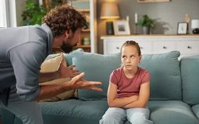 Ein Mann steht mit beiden Händen gestikulierend im Wohnzimmer vor einem auf dem Sofa sitzenden Mädchen. Das Mädchen hat die Arme vor dem Körper verschränkt und schaut ihm in die Augen.