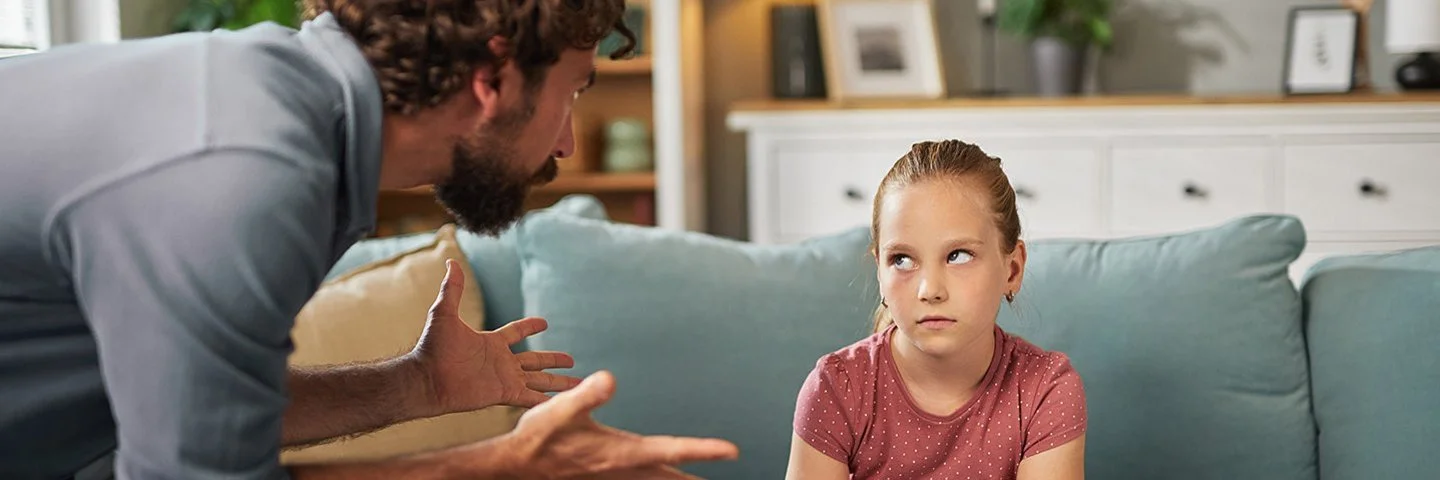 Ein Mann steht mit beiden Händen gestikulierend im Wohnzimmer vor einem auf dem Sofa sitzenden Mädchen. Das Mädchen hat die Arme vor dem Körper verschränkt und schaut ihm in die Augen.