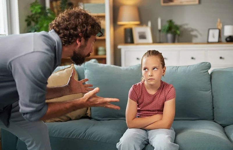 Ein Mann steht mit beiden Händen gestikulierend im Wohnzimmer vor einem auf dem Sofa sitzenden Mädchen. Das Mädchen hat die Arme vor dem Körper verschränkt und schaut ihm in die Augen.
