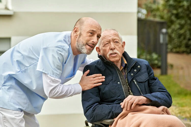 Eine Pflegeperson steht gebeugt neben einem älteren Mann, der in einem Rollstuhl sitzt.