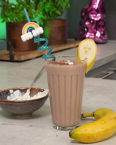 Schoko-Bananen-Proteinshake nach einem Rezept von Felicitas Then, angerichtet in einem hohen Glas mit buntem Strohhalm und Bananenscheibe am Rand. Rechts liegt eine Banane, links steht eine braune Schale mit Magerquark.