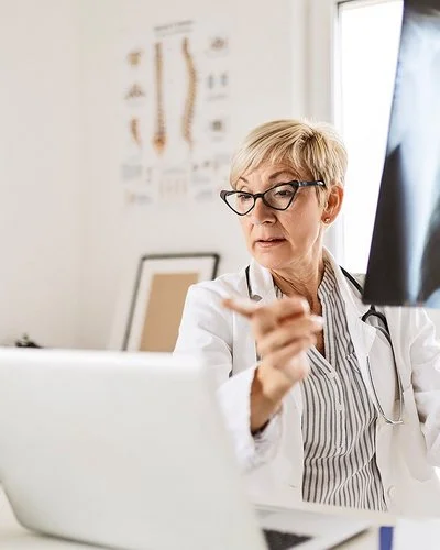 Eine Radiologin mittleren Alters und mit kurzen blonden Haaren sitzt am Schreibtisch vor ihrem Computer, in der linken Hand hält sie ein Röntgenbild eines Brustkorbs.