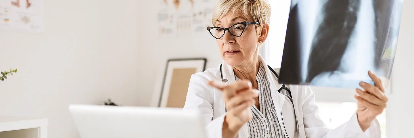 Eine Radiologin mittleren Alters und mit kurzen blonden Haaren sitzt am Schreibtisch vor ihrem Computer, in der linken Hand hält sie ein Röntgenbild eines Brustkorbs.