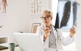Eine Radiologin mittleren Alters und mit kurzen blonden Haaren sitzt am Schreibtisch vor ihrem Computer, in der linken Hand hält sie ein Röntgenbild eines Brustkorbs.