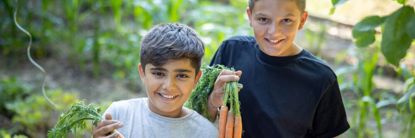 Zwei Schuljungen lächeln in die Kamera und halten frisch geerntete Karotten in den Händen.