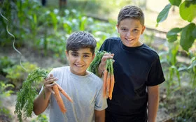 Zwei Schuljungen lächeln in die Kamera und halten frisch geerntete Karotten in den Händen.