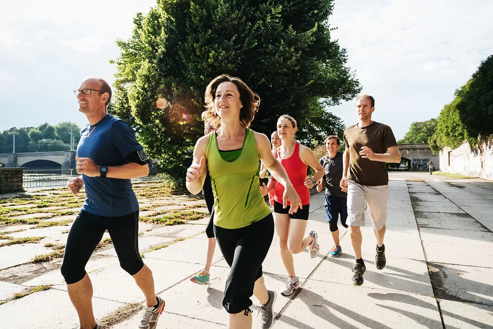 Eine Gruppe von Frauen und Männern joggt auf einem asphaltierten Weg durch einen Stadtpark. 