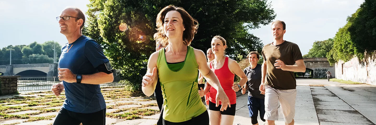 Eine Gruppe von Frauen und Männern joggt auf einem asphaltierten Weg durch einen Stadtpark. 