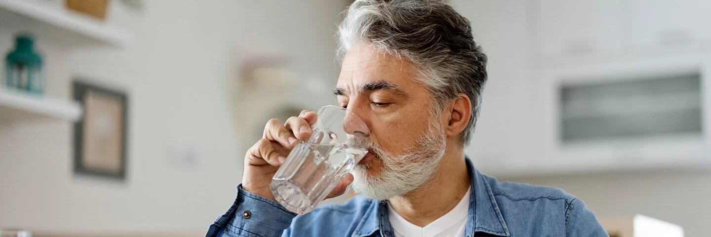 Gegen seine Mundtrockenheit trinkt ein älterer Mann Wasser aus einem Glas, während er am Küchentisch sitzt.