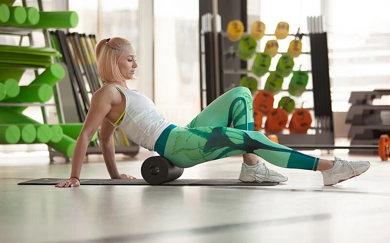 Eine junge Frau stützt sich neben der Gymnastikmatte mit den Händen ab. Das rechte Bein ist ausgestreckt, das linke aufgestellt. Mit einer Faszienrolle bearbeitet sie ihre Gesäßmuskeln.