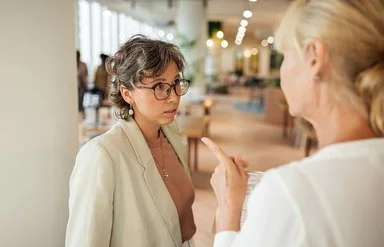 Eine Frau in Businesskleidung, die nur von hinten zu sehen ist, spricht mit einer anderen Frau und deutet mit dem Zeigefinger auf sie. Die andere Frau blickt leicht angespannt.