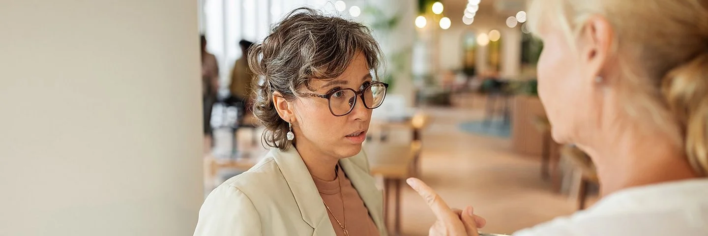 Eine Frau in Businesskleidung, die nur von hinten zu sehen ist, spricht mit einer anderen Frau und deutet mit dem Zeigefinger auf sie. Die andere Frau blickt leicht angespannt.