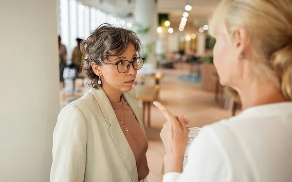 Eine Frau in Businesskleidung, die nur von hinten zu sehen ist, spricht mit einer anderen Frau und deutet mit dem Zeigefinger auf sie. Die andere Frau blickt leicht angespannt.