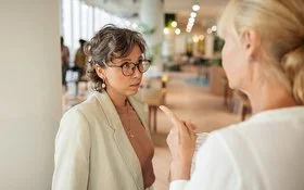 Eine Frau in Businesskleidung, die nur von hinten zu sehen ist, spricht mit einer anderen Frau und deutet mit dem Zeigefinger auf sie. Die andere Frau blickt leicht angespannt.