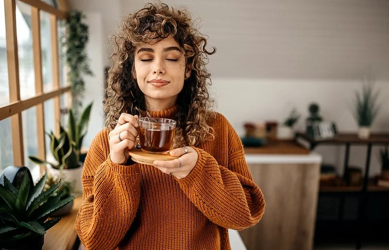 Eine Frau in einem braunen Pullover hält mit geschlossen Augen eine Tasse Tee in der Hand.