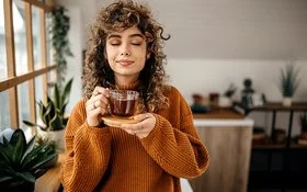 Eine Frau in einem braunen Pullover hält mit geschlossen Augen eine Tasse Tee in der Hand.